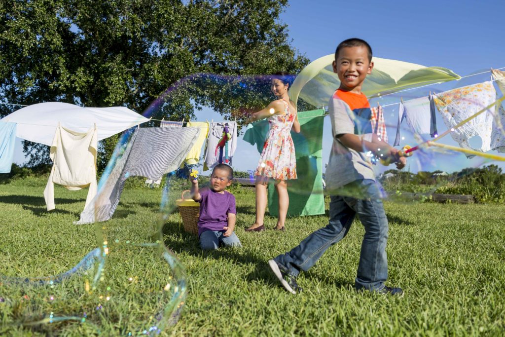 Two children play with bubbles while a young woman hangs laundry on an outdoor line. Amway Corporation and the Nutrilite brand are seriously committed to helping children grow up happy and healthy. 